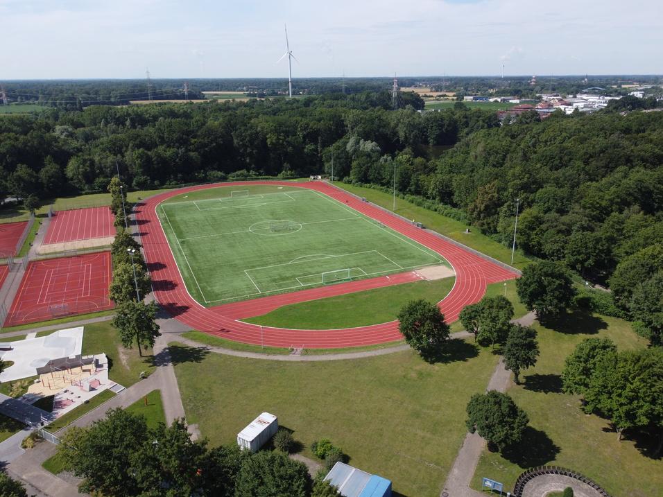 Luftaufnahme eines Sportgeländes mit einem Fußballfeld, Tennisplätzen und umliegendem Wald.