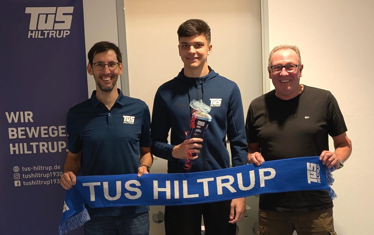Drei Personen stehen lächelnd zusammen, halten einen blauen Schal mit „TuS Hiltrup“ und eine Auszeichnung in der Hand.