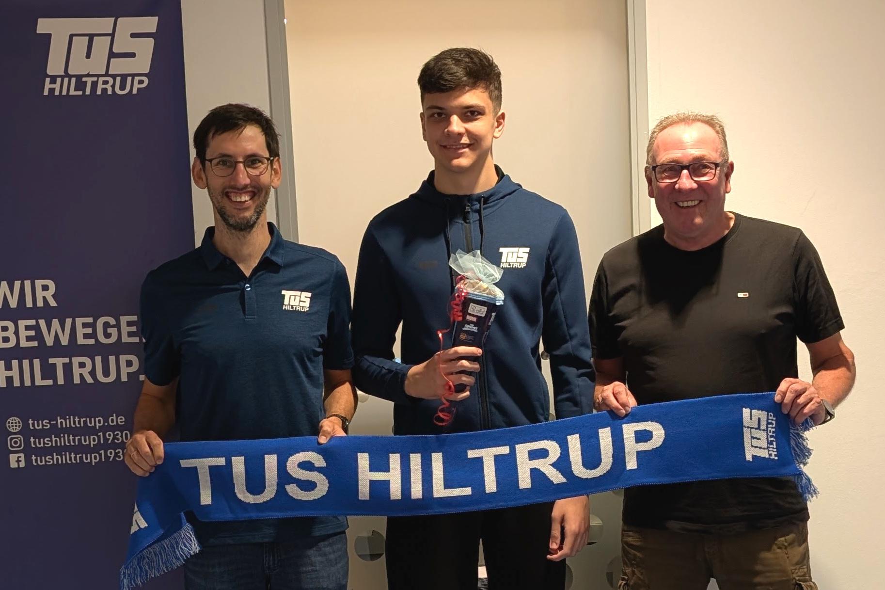 Drei Personen stehen lächelnd zusammen, halten einen blauen Schal mit „TuS Hiltrup“ und eine Auszeichnung in der Hand.