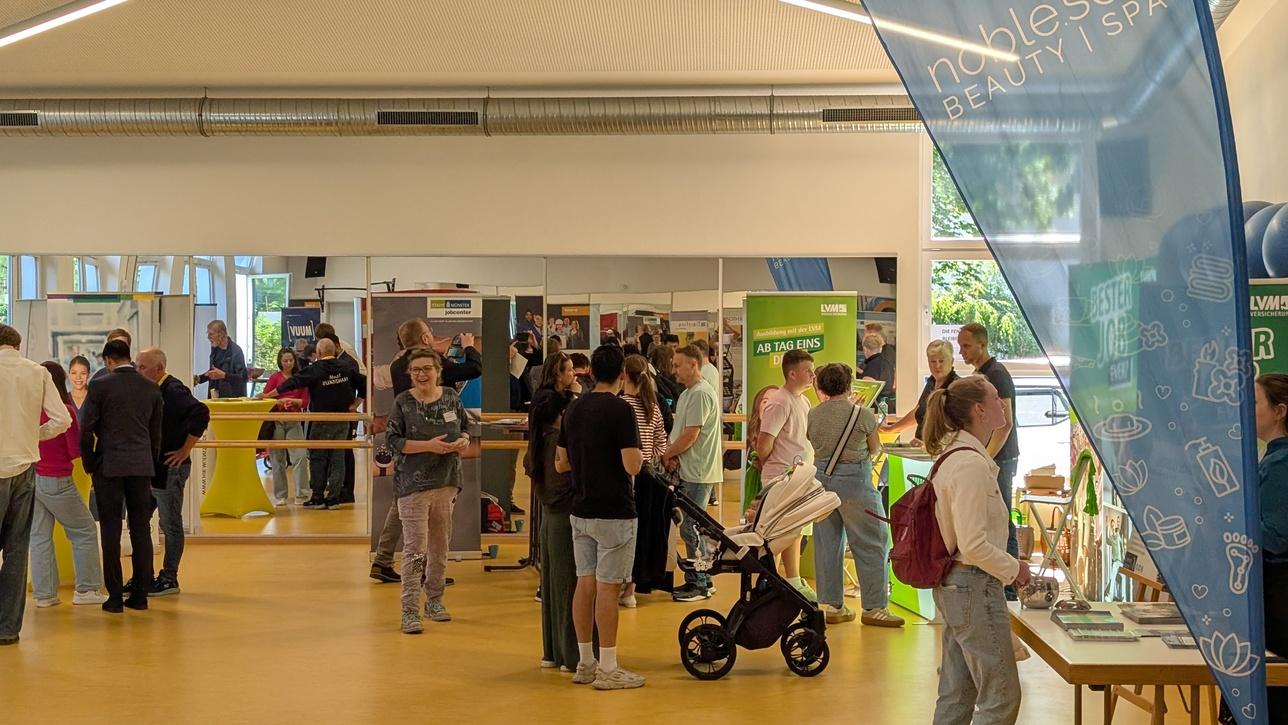 Messestand mit Menschen in einer großen Halle, Informationsstände und Banner im Hintergrund, helles und offenes Raumdesign.