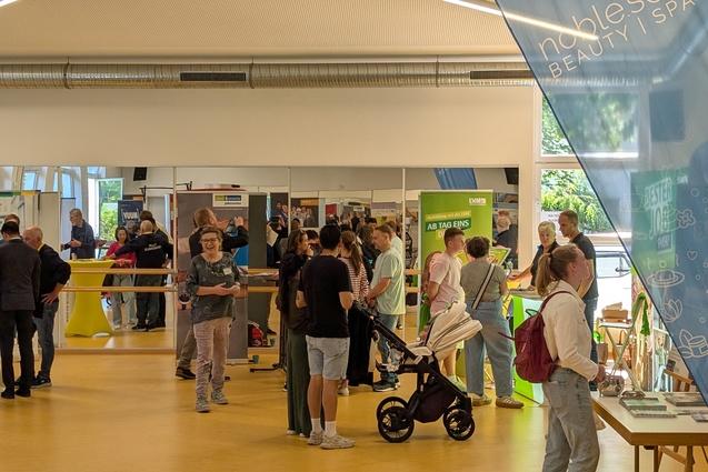 Messestand mit Menschen in einer gro&szlig;en Halle, Informationsst&auml;nde und Banner im Hintergrund, helles und offenes Raumdesign.