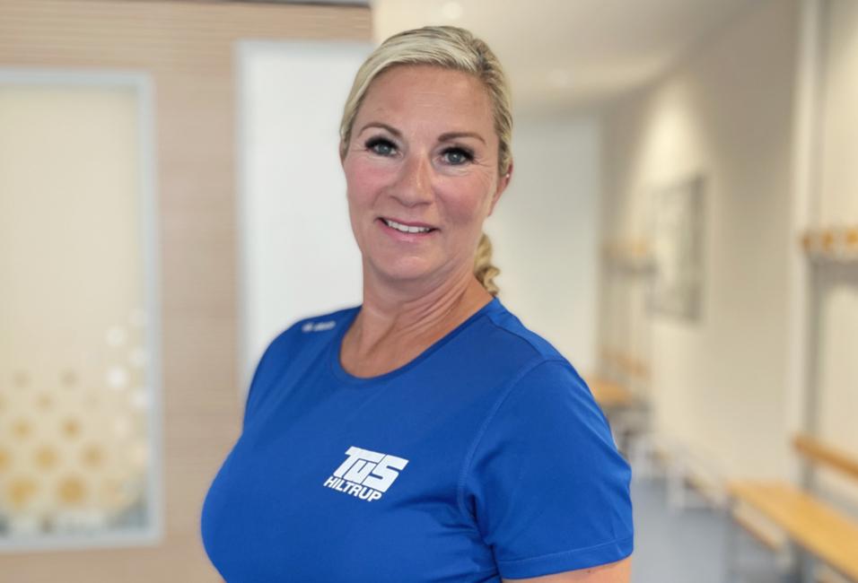 Frau mit blonden Haaren in blauem Shirt steht in einem hellen Raum mit Holzbänken im Hintergrund.