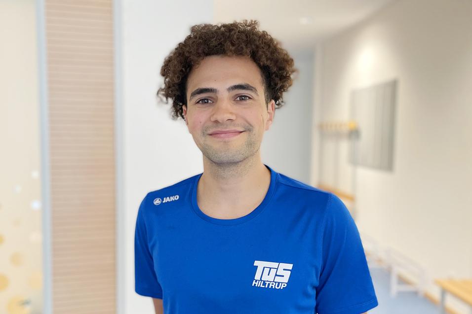 Junger Mann mit lockigem Haar in blauem Sportshirt l&auml;chelt vor einem hellen, modernen Hintergrund.