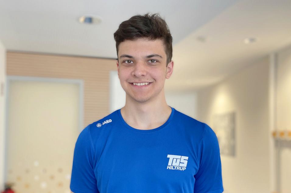 L&auml;chelnder junger Mann tr&auml;gt ein blaues T-Shirt mit dem Schriftzug &bdquo;TuS Hiltrup&ldquo;, Hintergrund unscharf mit hellem Raum.