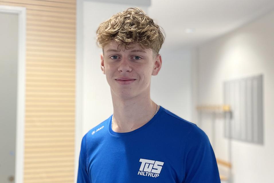 Junger Mann mit lockigem Haar tr&auml;gt ein blaues Sportshirt mit Aufschrift in einem hellen Innenraum.