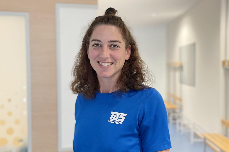 Sina Hofmayer Frau mit lockigem Haar in blauem Shirt steht in einem hellen Raum mit Holzbank im Hintergrund.
