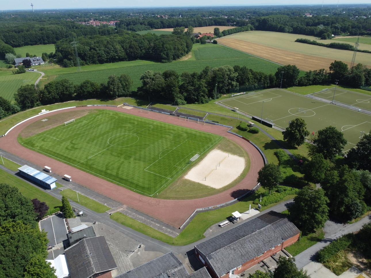 Luftaufnahme eines Fu&szlig;ballfeldes mit Leichtathletikbahn, Sandkasten und angrenzenden Sportpl&auml;tzen umgeben von Wald und Feldern.