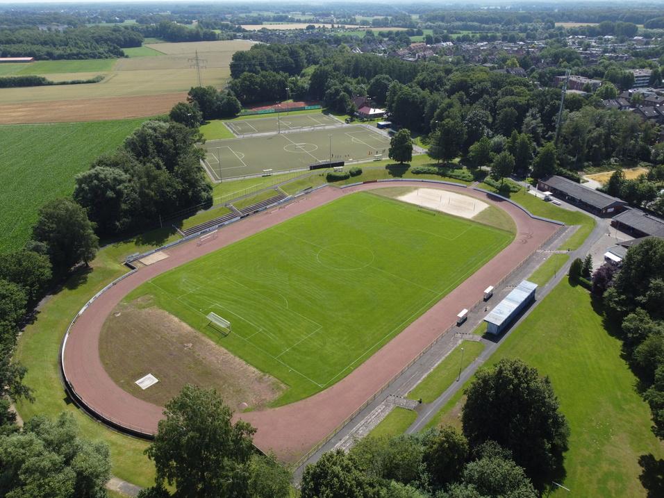 Luftaufnahme eines Sportgeländes mit einem Fußballfeld, einem Laufbahnring und einem angrenzenden Sandplatz.