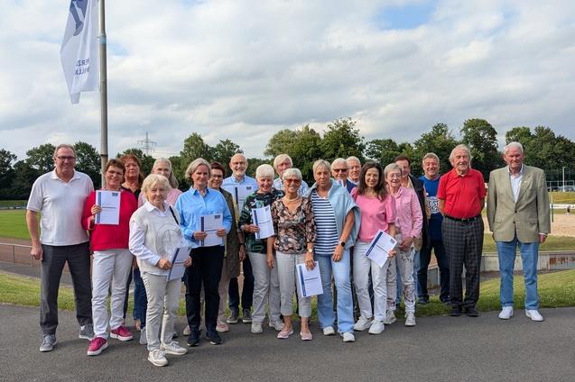 Gruppenfoto von 28 Menschen im Freien, die Urkunden in den Händen halten, vor einem Sportplatz mit Bäumen im Hintergrund.