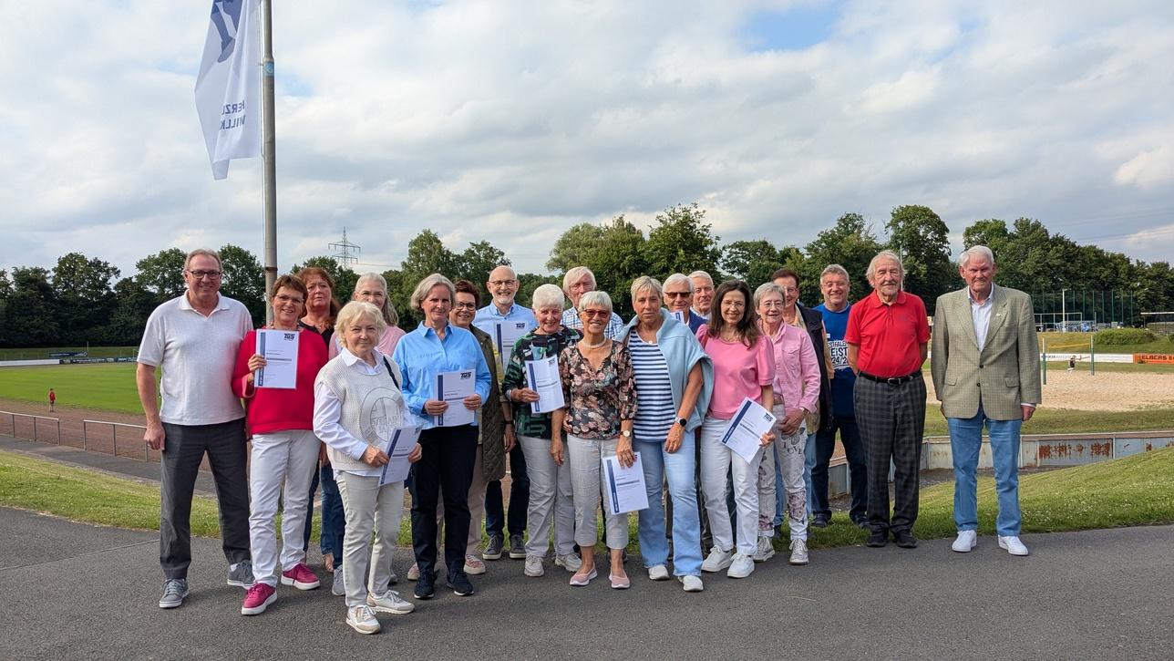 Gruppenfoto von 28 Menschen im Freien, die Urkunden in den Händen halten, vor einem Sportplatz mit Bäumen im Hintergrund.