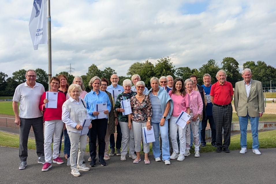 Gruppenfoto von 28 Menschen im Freien, die Urkunden in den Händen halten, vor einem Sportplatz mit Bäumen im Hintergrund.