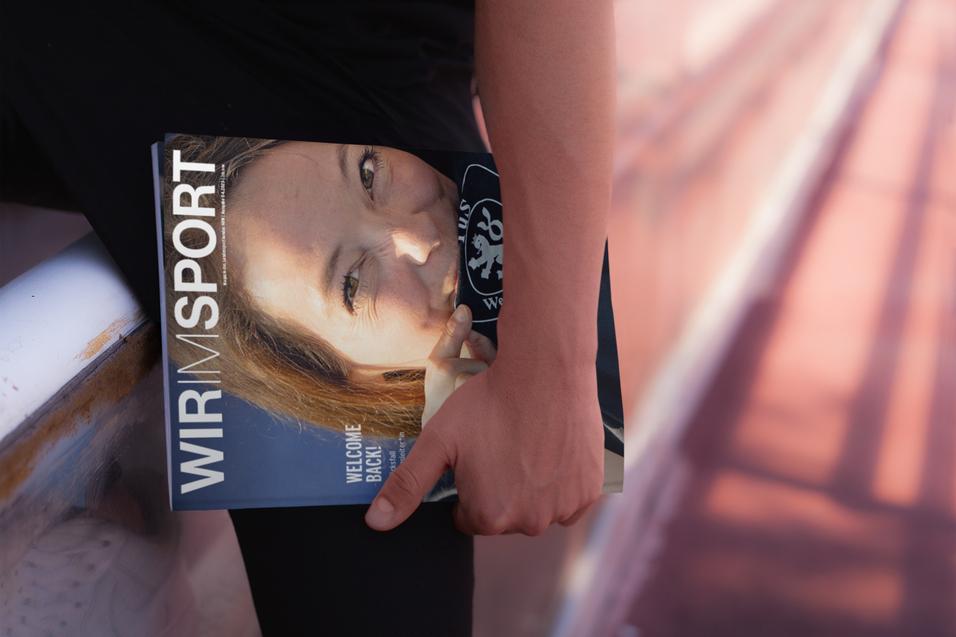 Nahaufnahme einer Hand, die ein Magazin mit dem Titel &bdquo;WIR IM SPORT&ldquo; h&auml;lt, Hintergrund in unscharfem Laufbahn-Design.