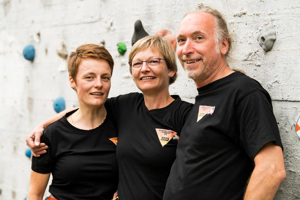 Drei Personen in schwarzen T-Shirts lächeln vor einer Kletterwand mit bunten Griffen.