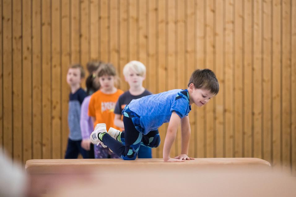 Ein Junge balanciert auf einem Blickkasten, während mehrere Kinder im Hintergrund zuschauen. Holzpaneele sind sichtbar.